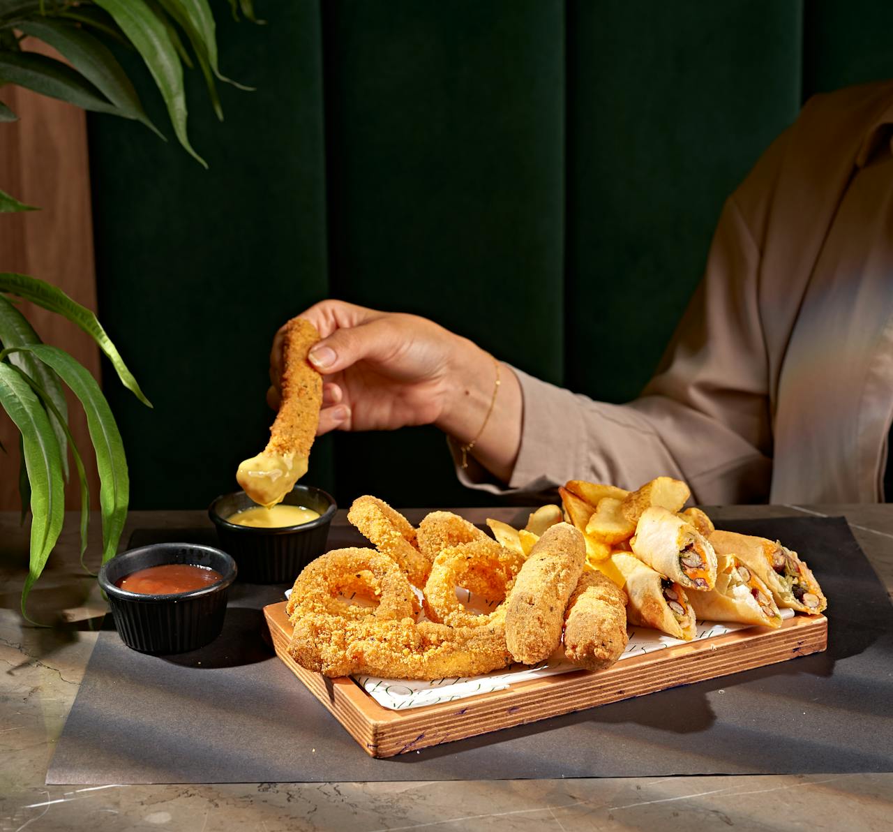 Hand dipping snack into sauce with assorted fried foods on platter.